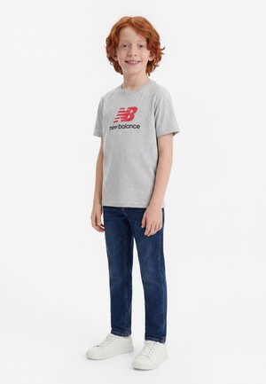 Un garçon roux souriant portant un t-shirt gris New Balance, un jean bleu et des baskets blanches, debout sur un fond blanc.