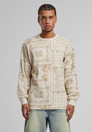 Beige long-sleeve shirt featuring a patchwork design with intricate brown patterns. Made of soft fabric, it has ribbed cuffs and a crew neckline.