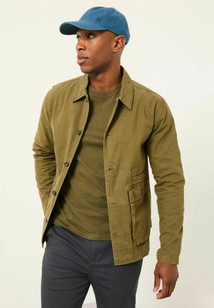 Olivgrüne Baumwolljacke mit Frontknöpfen und Seitentaschen, getragen über einem dunkelgrünen Hemd, kombiniert mit einer blauen Mütze und dunklen Hosen.