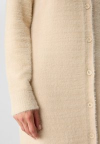Cardigan beige in un tessuto texturizzato e fuzzy, con una chiusura con bottoni e polsini a costine. Presenta tonalità morbide e neutre in tutto il capo.