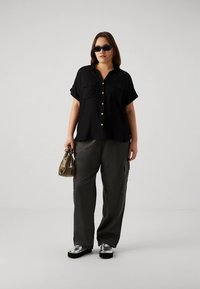 Črna kratka srajca s kratkimi rokavi in zlatimi gumbi, kombinirana s sivimi hlačami na žepe in srebrnimi platformnimi čevlji. Dodatek: majhna bež torbica.