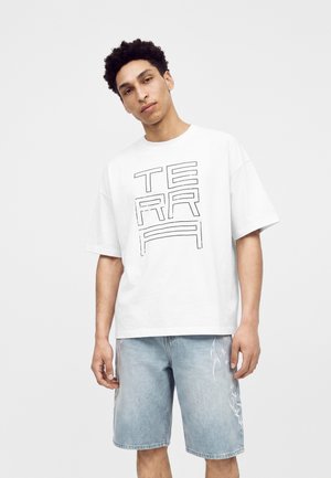 Mladý muž nosiaci biele oversized tričko s textom "TERRA" a svetlomodré rifľové šortky s bielymi plameňovými vzormi.