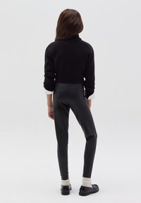 Suéter negro de punto con cuello alto, combinado con leggings negros brillantes. La modelo lleva puños blancos y zapatos negros con suelas gruesas. Texturas suaves.