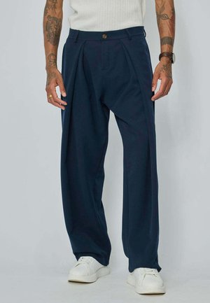 Homme portant un pantalon plissé ample bleu foncé, des baskets blanches, un top blanc sans manches, avec des bras tatoués et une montre-bracelet.
