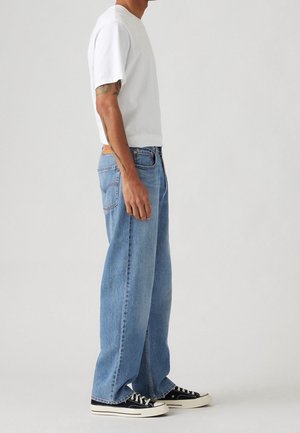 Mann trägt ein weißes T-Shirt mit kurzen Ärmeln, hellblaue locker sitzende Jeans, schwarze niedrige Sneakers, steht im Profil vor schlichtem grauem Hintergrund.
