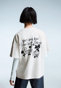 Crème katoenen t-shirt met korte mouwen, voorzien van zwarte tekst en bloemgrafieken op de rug, gecombineerd met donkere spijkerbroek.