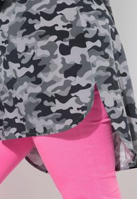Grå camouflage-mønstrede top med en buet kant og sidesplitter, parret med lyserøde tætsiddende bukser. Begge dele har en glat tekstur.