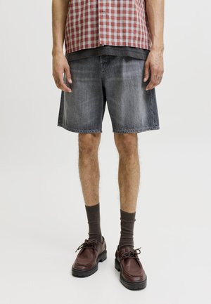 Homme portant un short en denim noir délavé, des chaussettes marron et des chaussures en cuir marron foncé, debout devant un fond uni.
