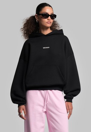 Sort oversized hættebluse med en frontlomme og hvid tekst "WRSTBHV" på brystet, parret med lyserøde sweatpants. Glat, blød tekstur.