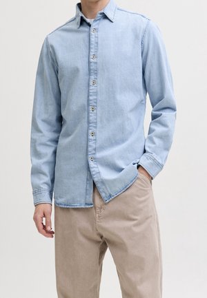 Mężczyzna w jasnoniebieskiej, zapinanej na guziki koszuli jeansowej i beżowych spodniach, z jedną ręką w kieszeni i neutralnym tłem.