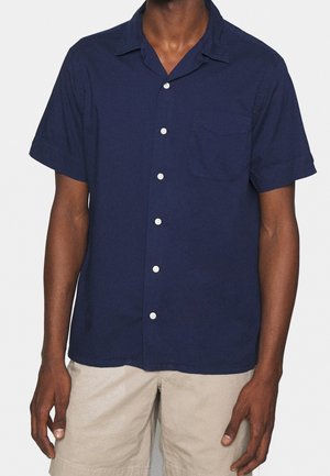 Homme portant une chemise bleu marine à manches courtes avec col et un short beige clair sur un fond uni.