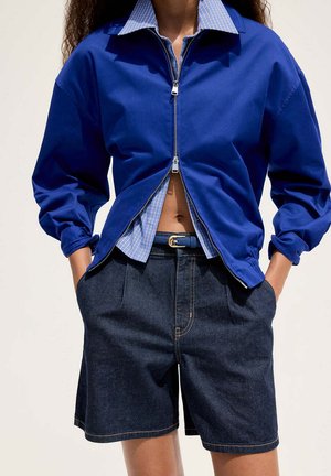 Femme portant une veste bleue à fermeture éclair sur une chemise à carreaux et un short bermuda en jean foncé, les mains dans les poches, sur un fond neutre.