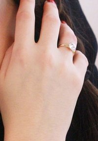 Anello d'oro con una pietra preziosa trasparente a forma di pera, indossato su una mano con pelle chiara e smalto per unghie, su uno sfondo scuro.