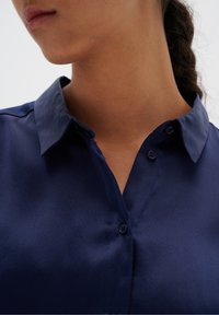Blusa di satin blu navy con colletto a punta, chiusura con bottoni e texture liscia. Dettaglio del colletto e dei bottoni visibile.