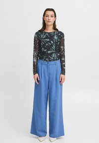 Schwarzes Langarmoberteil mit türkisfarbenem Batikmuster, kombiniert mit hellblauen weit geschnittenen Hosen. Das Model trägt weiße spitze Schuhe.
