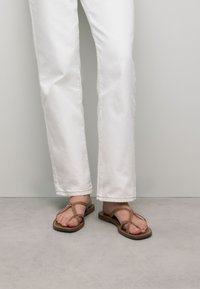 Bruine platte sandalen met kruislings gespen, platte zool en minimale details, gedragen met witte rechte broek tegen een neutrale achtergrond.
