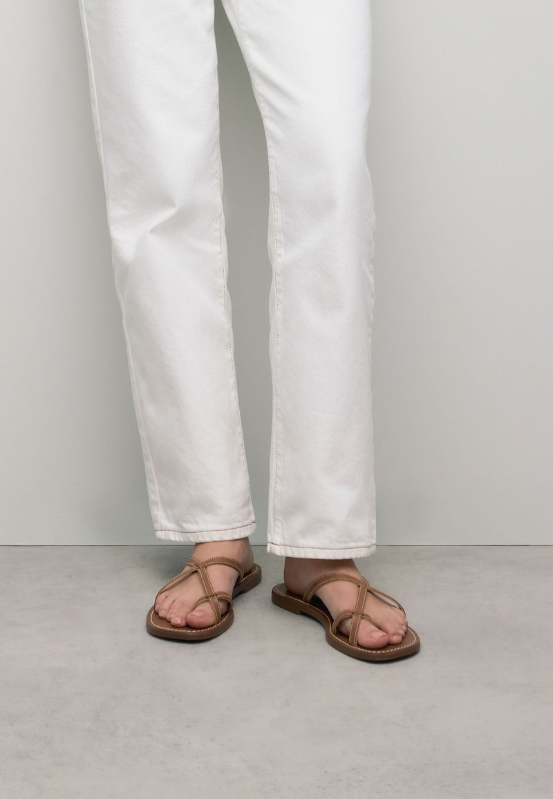 Bruine platte sandalen met kruislings gespen, platte zool en minimale details, gedragen met witte rechte broek tegen een neutrale achtergrond.