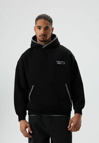 Zwarte hoodie van een katoenmengsel met een kangoeroezak, contrasterende witte ritsaccenten en wit geborduurde tekst op de borst.