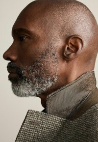 Profilul unui bărbat cu păr scurt și barbă gri, purtând o jachetă maro texturată cu guler cu model.