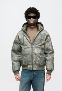 Prešívaná bunda s maskáčovým vzorom v tlmených odtieňoch zelenej a šedej, s predným zipsom, kapucňou a elastickými manžetami. Nosená cez hnedú košeľu.