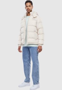Lys beige puffjakke med lynlås, hætte og broderet logo, båret over en lys blå skjorte og parret med blå jeans og hvide sneakers.