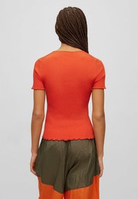 Rød ribbet T-shirt med korte ærmer, tætsiddende form, bølget kant og rund halsudskæring, parret med løse bukser i flere farver i grøn og orange.