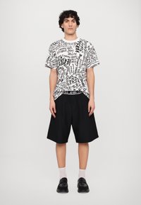 Melnbalts grafiska t-krekls ar dažādiem tekstu rakstiem, apvienots ar brīvi pieguļošām melnām šortiem. Modelis valkā melnas kurpes un baltas zeķes.