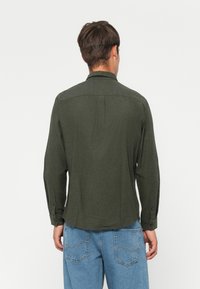 Uomo di spalle che indossa una maglietta a maniche lunghe verde scuro e jeans azzurri chiari su uno sfondo bianco uniforme.
