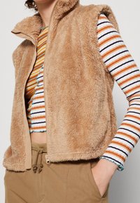 Tan faux päls väst med hög krage och dragkedja, med sidosyft och mjuk textur, i kombination med en flerfärgad randig långärmad skjorta.