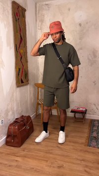 Νεαρός άνδρας με πράσινα σορτς και μπλουζάκι, με πορτοκαλί καπέλο τύπου κάσκα, μαύρη τσάντα χιαστί, λευκά αθλητικά παπούτσια, στέκεται μέσα σε κλειστό χώρο δίπλα σε καρέκλα και καφέ δερμάτινη τσάντα.