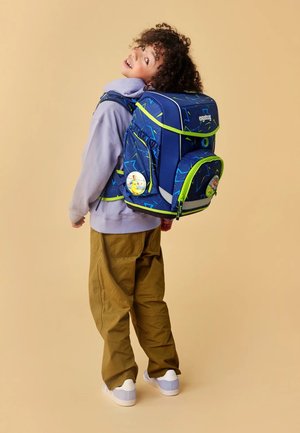 Mittelgroßer blauer Rucksack mit grünen Akzenten, geometrischen Mustern und reflektierenden Details; getragen von einem Kind in lässiger Kleidung.