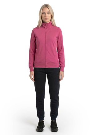 Une femme porte une veste rose zippée, un pantalon de jogging noir et des baskets noires sur un fond blanc uni.