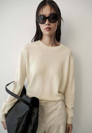 Mujer con gafas de sol negras de gran tamaño, suéter crema y pantalones beige, sosteniendo un gran bolso de cuero negro contra una pared lisa.