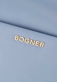 Un tessuto azzurro chiaro con texture presenta un logo dorato "BOGNER" ben in vista al centro. Il materiale ha un sottile motivo a trama.