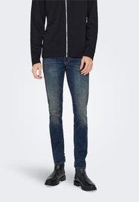 Tumši zilas džinsa bikses ar izbalējušu tekstūru, apvienotas ar melnu rāvējslēdzēja jaku un melniem potīšu zābakiem, radot gludu un plūstošu izskatu.