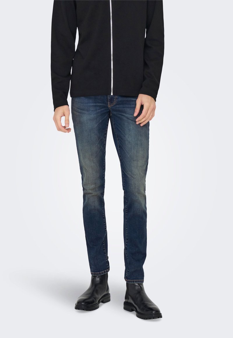 Tumši zilas džinsa bikses ar izbalējušu tekstūru, apvienotas ar melnu rāvējslēdzēja jaku un melniem potīšu zābakiem, radot gludu un plūstošu izskatu.