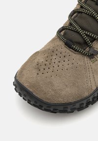 Zapato de ante marrón con parte superior texturizada, suela de goma negra y cordones con patrón. Presenta perforaciones cerca de la puntera para transpirabilidad.