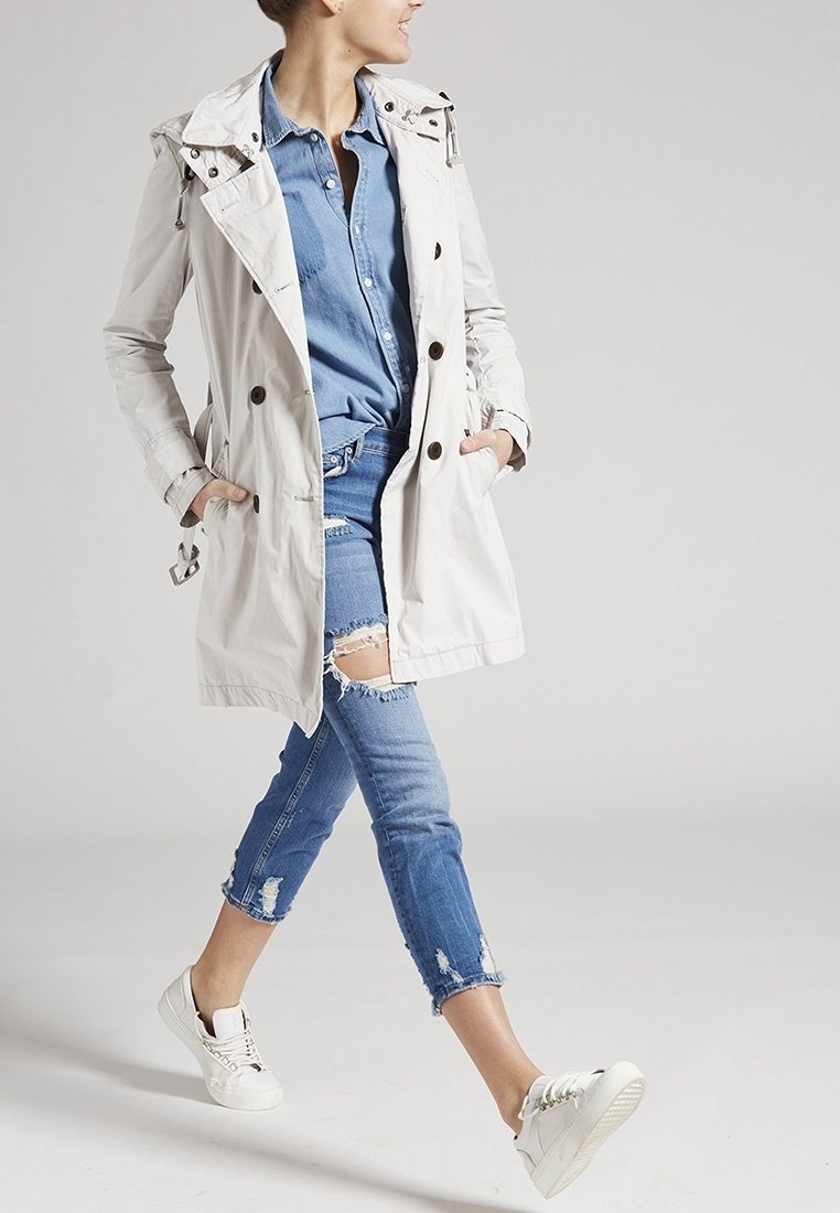 Trinchera ligera de crema con botones de doble pecho, llevada sobre una camisa de mezclilla azul y jeans desgastados. Zapatillas blancas completan el look.