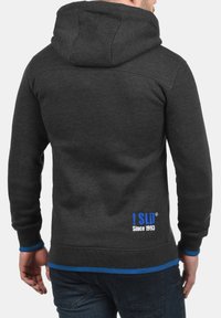 Sudadera con capucha de color gris oscuro, con una tela texturizada, que presenta un acento azul en el dobladillo y un logo con "Desde 1993" en la parte posterior. Diseño con capucha.