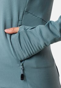 Teal long-sleeve top with zippered side pocket, ribbed cuffs, and smooth, stretchy fabric. Close-up of fabric texture and pocket detail.
