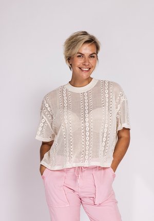 Smiling woman with short blonde hair wears a white crochet short-sleeve top and light pink pants with hands in pockets against a plain background.