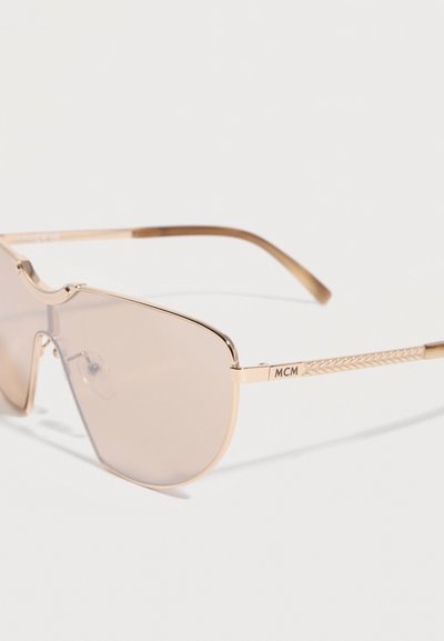 Γυαλιά ηλίου με χρυσή κορνίζα, με ένα κυρτό φακό σε ανοιχτό καφέ χρώμα και διακοσμητικούς βραχίονες, που φέρουν την επωνυμία "MCM" στο μπράτσο.