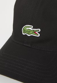 Casquette noire avec une surface texturée, arborant un logo de crocodile vert brodé avec un détail rouge sur le côté.