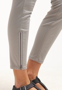 Pantaloni grigi chiaro con un design aderente, dotati di una cerniera laterale sulla caviglia. Tessuto di consistenza liscia e lunghezza cropped su misura.