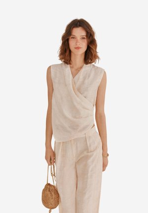 Haut sans manches en lin beige avec un design drapé, accompagné de pantalons larges assortis. Le modèle tient un petit sac tissé.