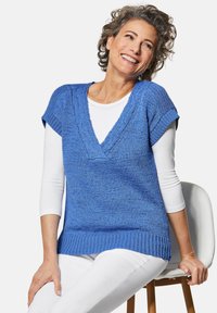 Blauw gebreid t-shirt met korte mouwen en een V-hals, met ribbelaccenten en een textuurafwerking, gedragen over een witte blouse met lange mouwen.