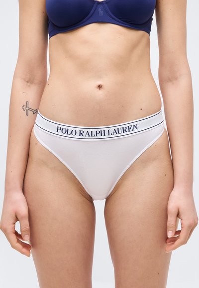 Γυναίκα που φοράει σουτιέν σε ναυτικό μπλε και λευκά εσώρουχα με το λογότυπο Polo Ralph Lauren, στέκεται με τα χέρια χαλαρά, δείχνοντας ένα τατουάζ στον αριστερό πήχυ.