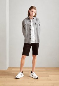 Camisa de botones gris con dos bolsillos en el pecho, camiseta blanca, pantalones cortos negros y zapatillas blancas con detalles negros, posando sobre un suelo de madera.