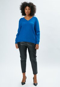 Blauer gestrickter Pullover mit V-Ausschnitt, kombiniert mit schwarzen, taillierten Pants aus Kunstleder und schwarzen High Heels. Glatte Textur, lässige Passform.