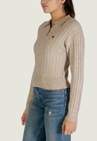 Beige kabelgebreide trui met een kraag, geribbelde manchetten en zoom. Voorzien van een kleine rode hartborduur op de borst. Gecombineerd met een spijkerbroek.